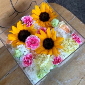 アクリルケースに収められた向日葵の花とピンクと黄色のお別れ用の花々。